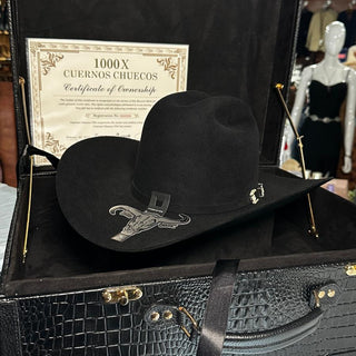 A black cowboy hat with silver embroidery rests inside an open black crocodile-embossed presentation case. A certificate is visible inside the case, and the hat has small silver decorative details.
