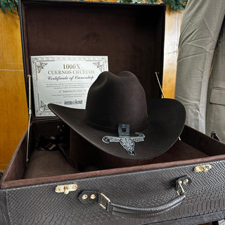 A dark brown cowboy hat with a silver skull emblem sits inside an open textured brown suitcase, with a certificate card visible in the lining. A person in a beige suit is partially visible on the right, and stacked boxes are in the background.