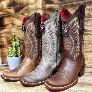 Three pairs of Los Altos Women’s Dubai Western Boots in varying shades of brown displayed on a wooden background with a cactus beside them.