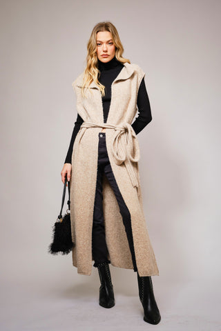 full view of long beige knit vest with waist tie over a black turtleneck and pants, styled in modern western fashion