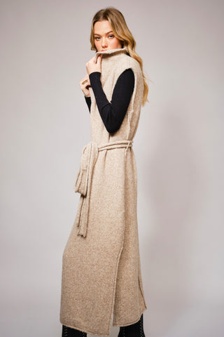side view of long beige knit vest with waist tie over a black turtleneck and pants, styled in modern western fashion