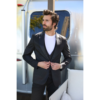 A man wearing a Black patterned Blazer with matching vest under with a white t-shirt, standing in front of a silver trailer.