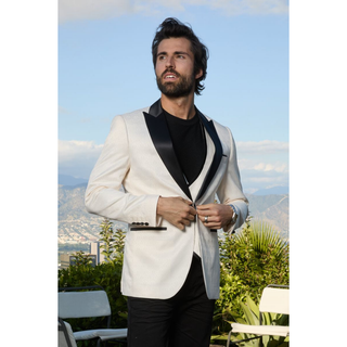 A man wearing a white blazer with black satin lapels, a black shirt, and black trousers, standing outdoors with a scenic mountain background.
