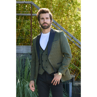 A man wearing an olive green patterned blazer with black satin lapels, matching vest, a white t-shirt, and black trousers, standing outdoors in front of green plants.