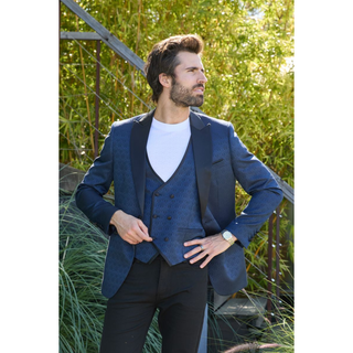 A man wearing a blue patterned blazer with a matching vest underneath, standing outdoors in front of green plants.
