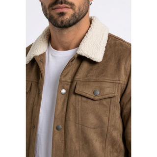 A close-up of a man wearing a brown faux suede jacket with a cream sherpa collar over a white t-shirt, against a plain white background.