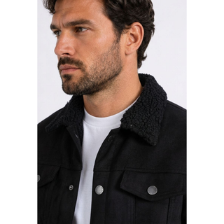 A close-up of a man wearing a black faux suede jacket with a plush black sherpa collar over a white t-shirt, shown from the chest up against a light gray background.